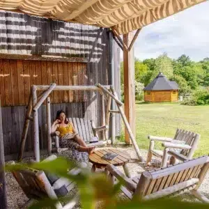Mobiliers pour extérieur, en bois de cèdre blanc massif du Canada. Représentation de l'hébergement insolite Ecôtelia, qui pose avec la balancelle en bois blanc naturel et un ensemble de fauteuils et canapé en bois canadien, tendance et sans traitement.